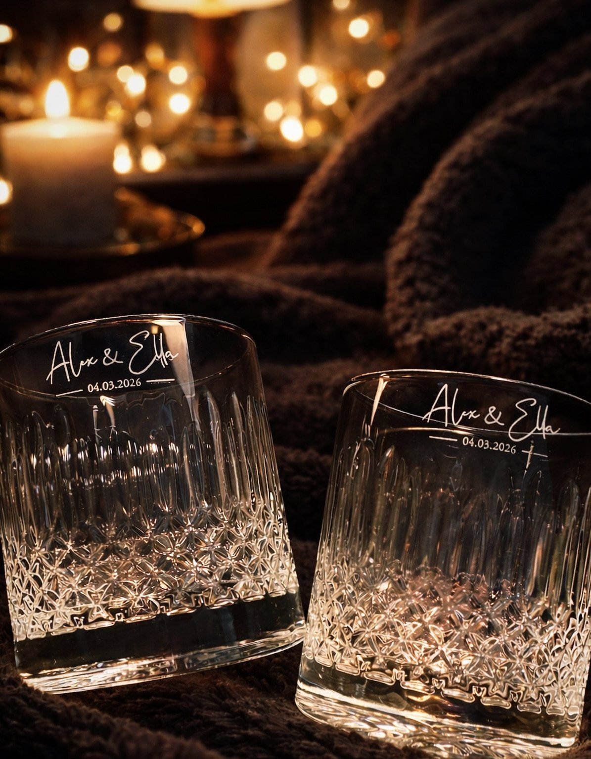 Two personalized crystal whiskey glasses on a brown blanket with warm bokeh lights.