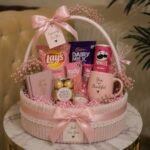 Pink themed gift basket with snacks, a mug, and flowers on a marble table.