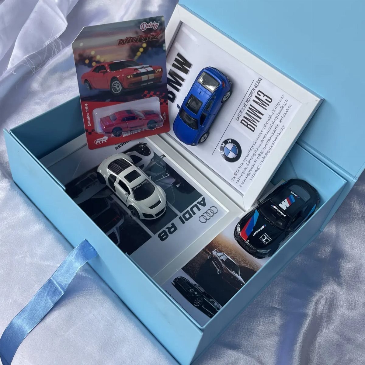 Miniature toy cars in a light blue gift box on white fabric.