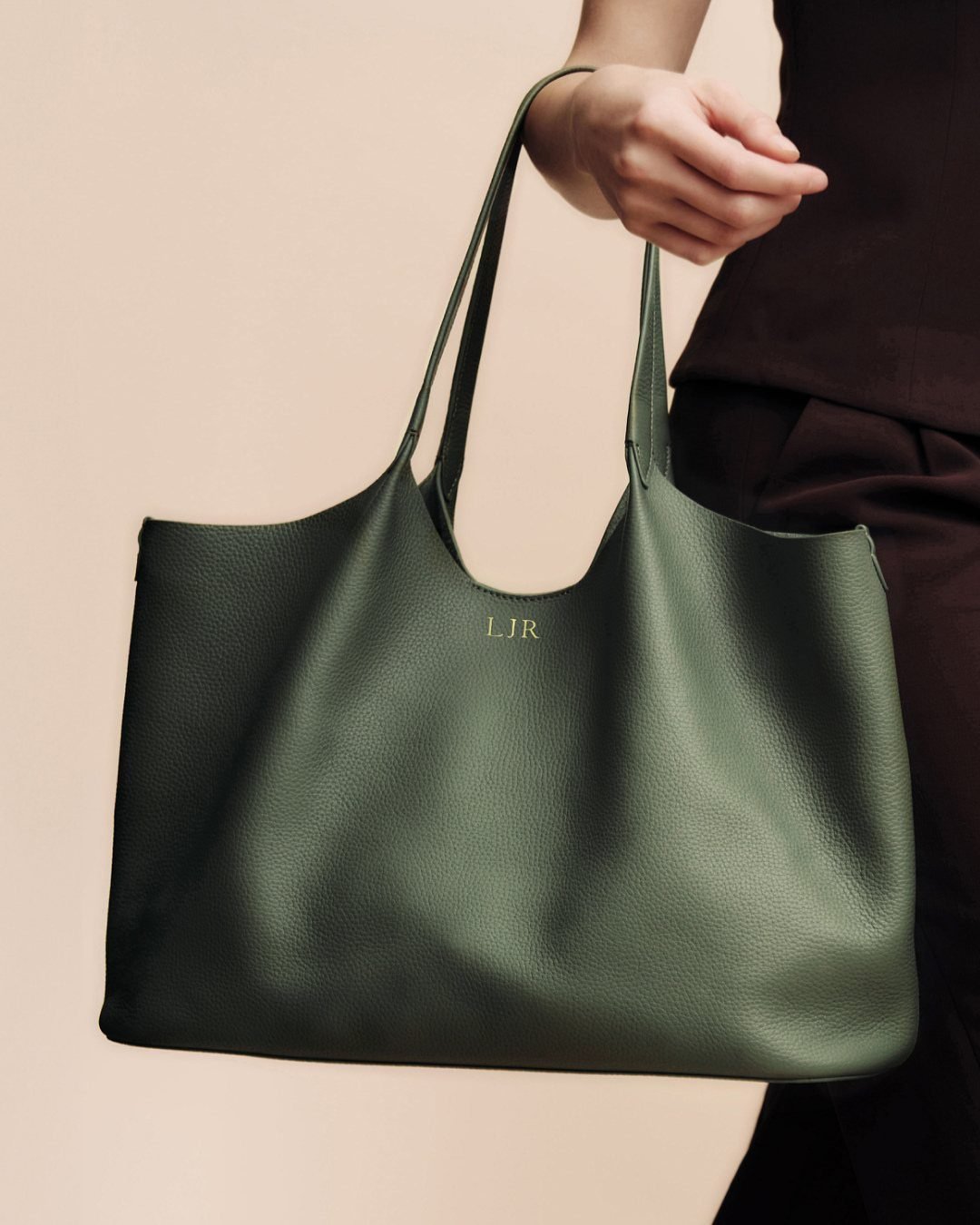Dark green pebbled leather tote bag with gold embossed initials held by person