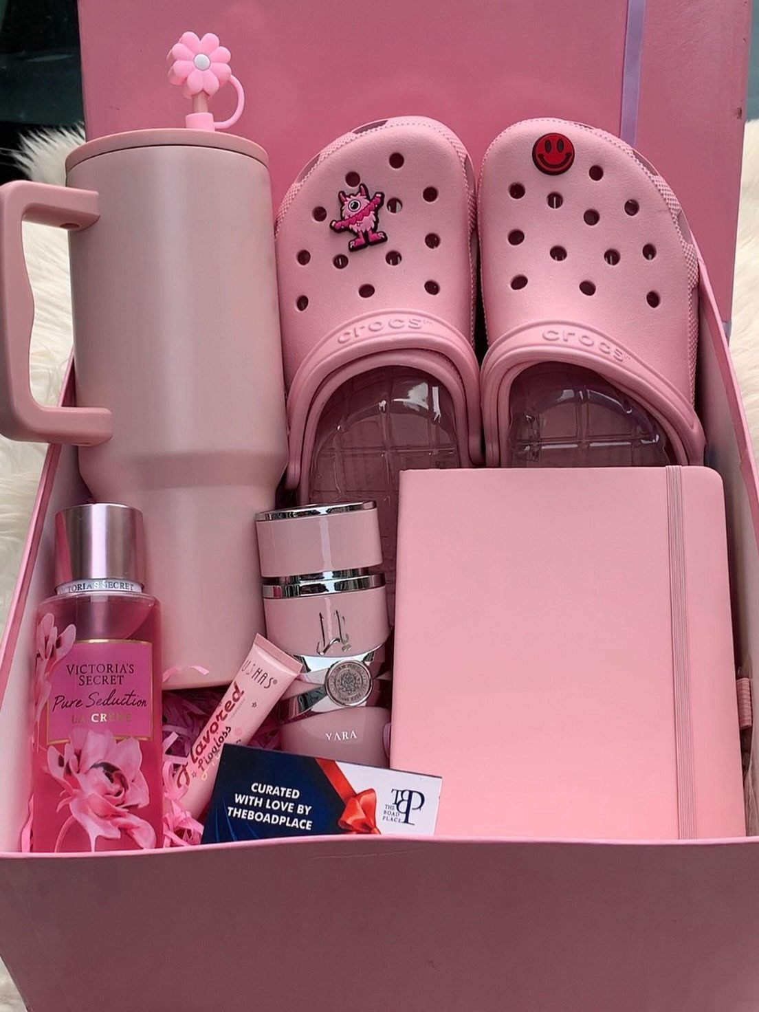 Pink gift box filled with a tumbler, Crocs, notebook, and perfume for Valentine's Day.