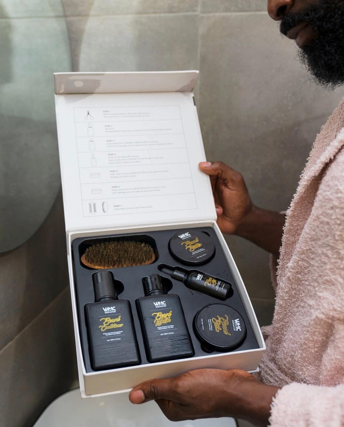 Man holding an open white box containing a complete beard grooming kit.